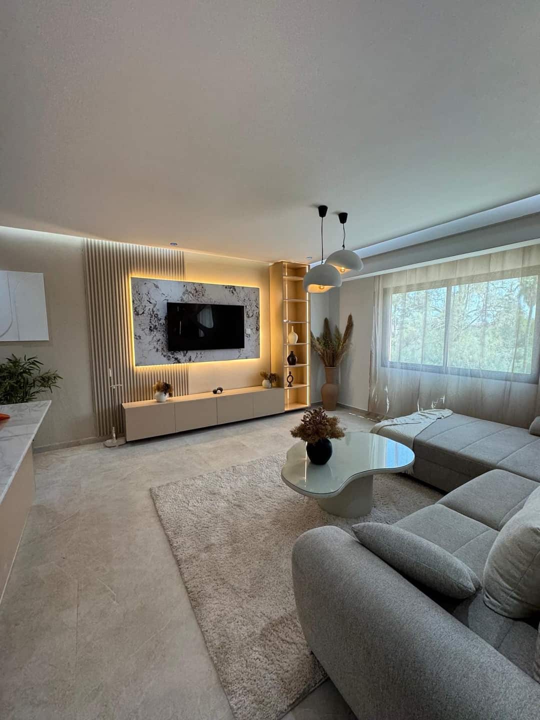 Séjour avec canapé d'angle, table basse blanche, mur TV rétroéclairé et fenêtre à voilages